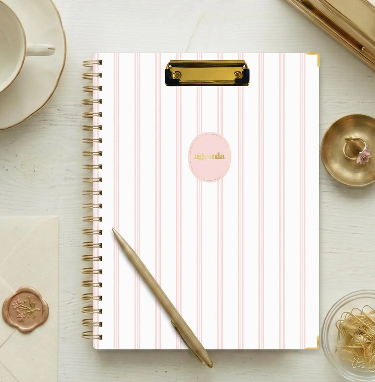 A spiral-bound non-dated planner with a white cover featuring vertical light pink stripes, a gold clip at the top, and a pink oval label in the center with the word &