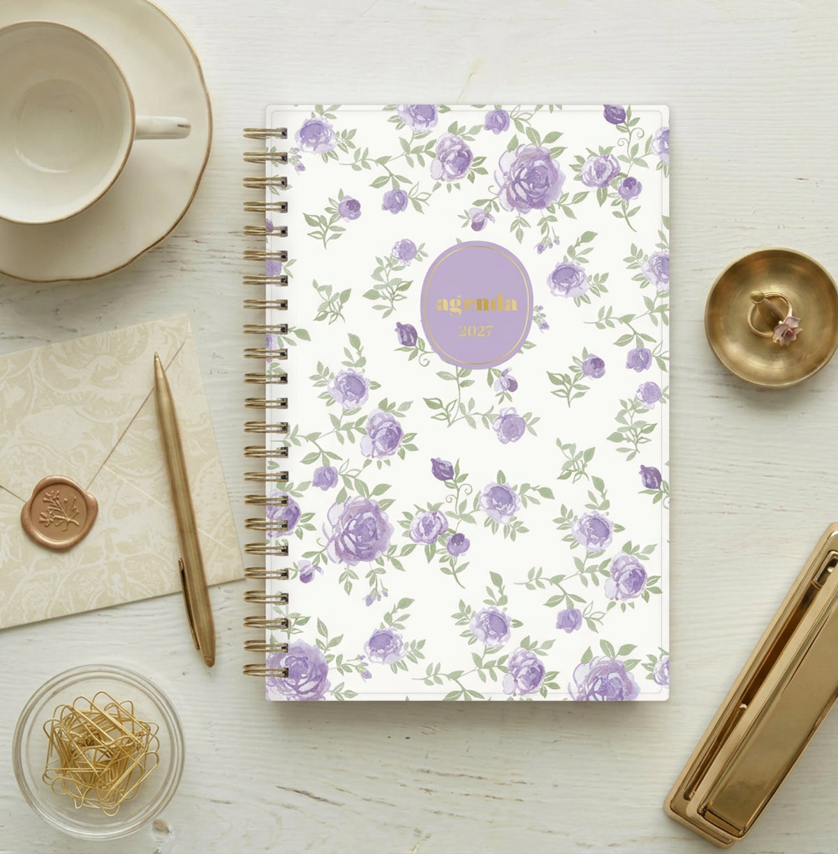 A spiral-bound planner agenda with a white cover decorated with a purple rose and green leaf pattern, featuring a lavender oval label with gold foil text that reads &