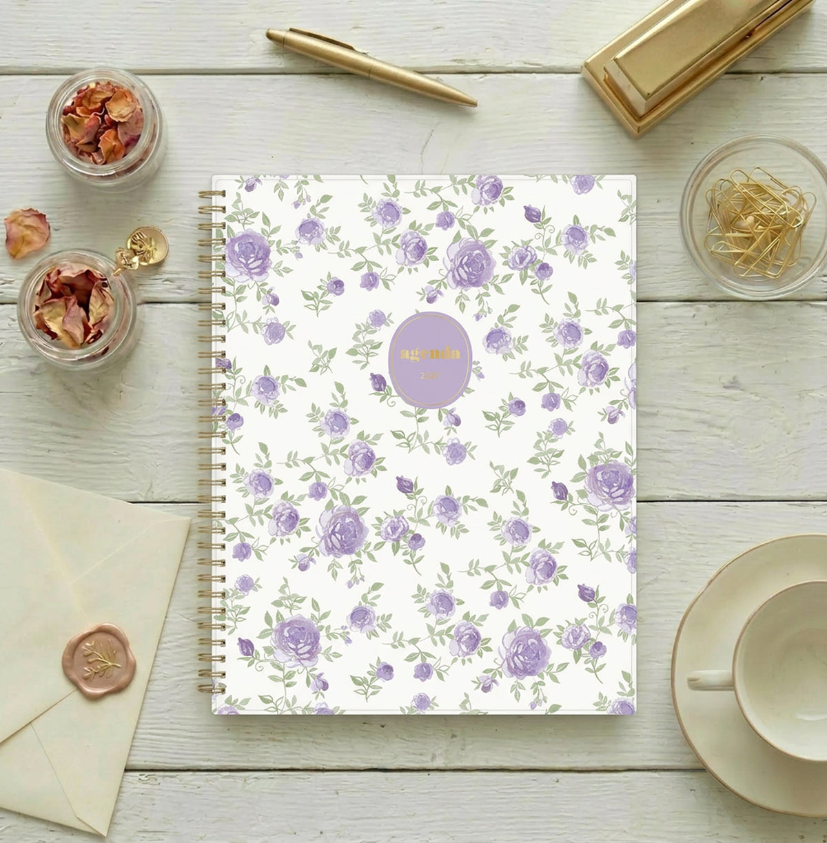 A spiral-bound weekly monthly planner with a white cover decorated with a repeating pattern of purple roses and green leaves. A lavender circular sticker with gold foil text reading &