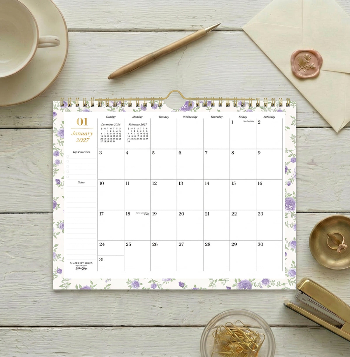 A spiral-bound monthly calendar page for January 2027, featuring a white background with light purple floral borders and gold accents, including a gold spiral binding and a gold clip at the top.