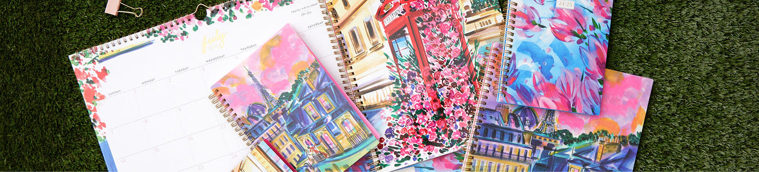 A spread of vibrant, colorful Travel Write Draw calendars and spiral-bound planners laid across green turf