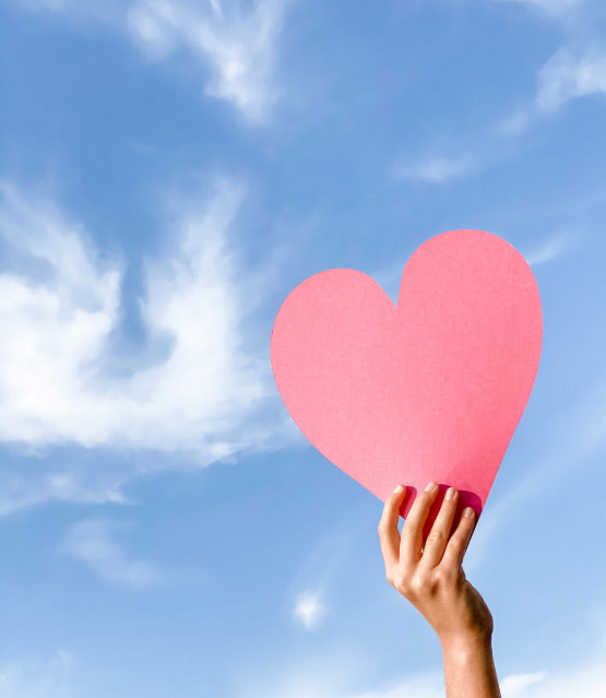 A hand holding up a red paper heart against a blue sky