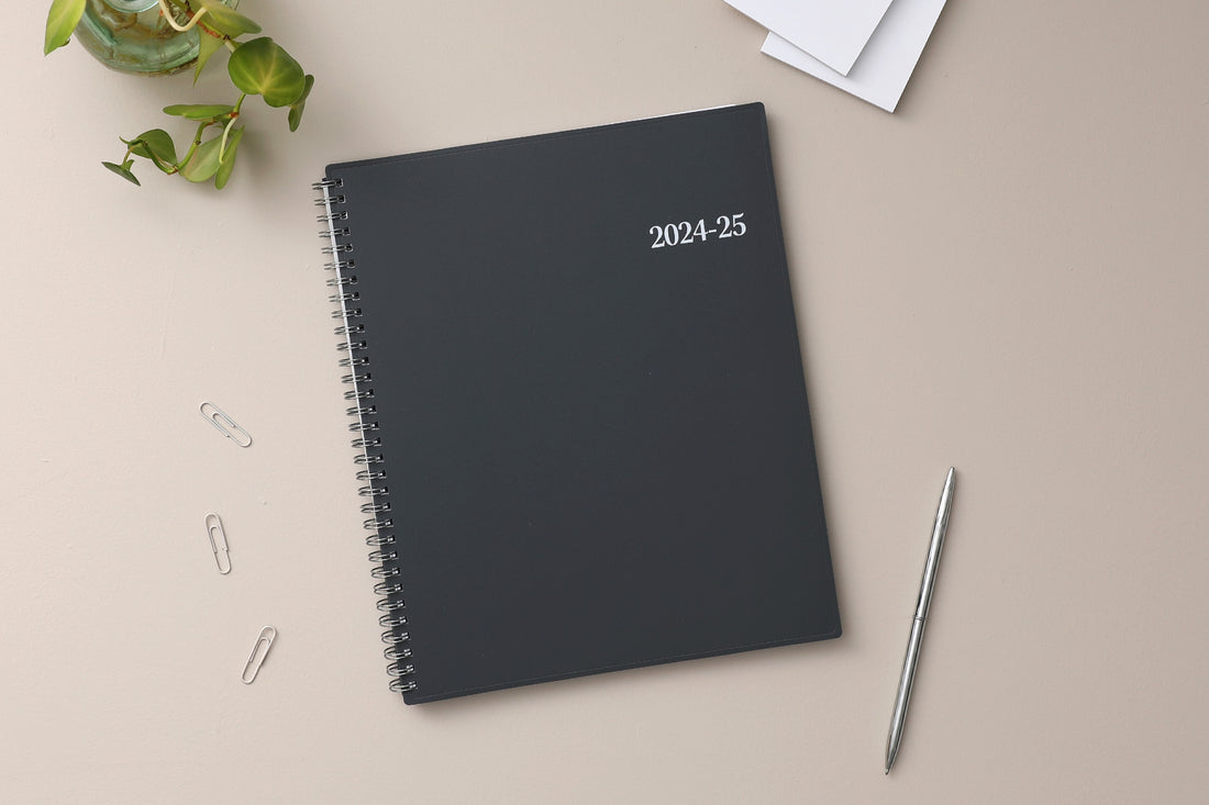 June 2024 weekly monthly academic planner featuring a solid charcoal cover and silver twin wire-o binding 8.5x11 size on a beige background, with a pen, paper clips, notes pages, and plant for lifestyle photo.