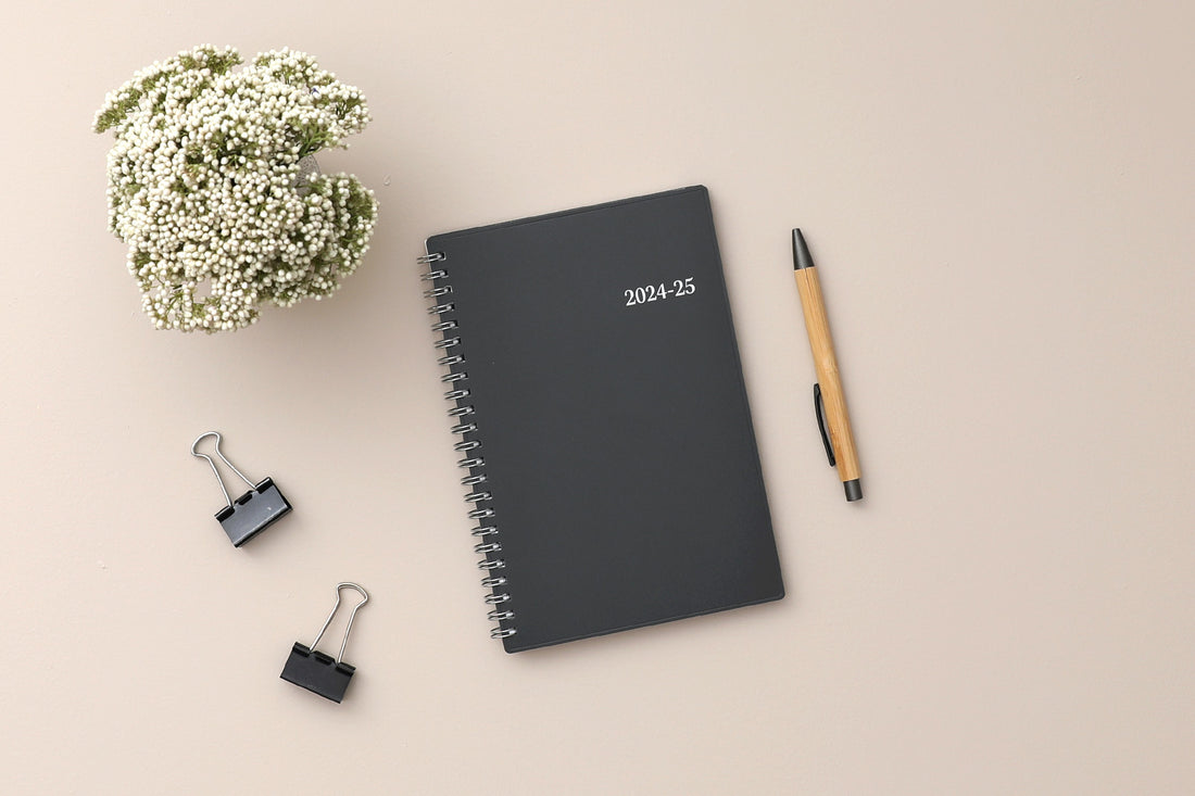monthly academic professional planner featuring a solid charcoal cover and silver twin wire-o binding in 8.5x11 placed on a beige background, pen, paperclip, and plant lifestyle photo