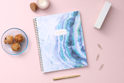 weekly monthly academic planner featuring a geo crystal front cover with gold twin wire-o binding in 8.5 x 11 planner size for 2024-2025 academic year, lifestyle photo with stapler, paperclip, muffins, candle, and pen on the side
