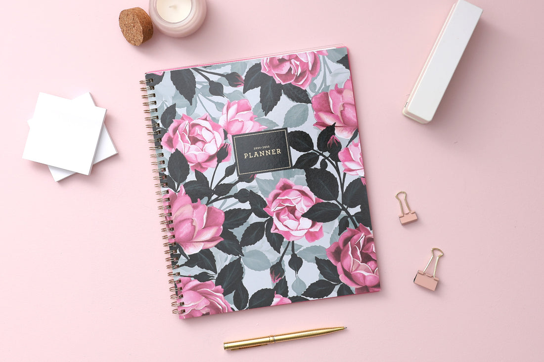 weekly monthly academic planner featuring a pink roses, shaded rose pedals, gold twin wire-o binding, and a compact 8.5x11 planner size. Lifestyle photo on pink background, paperclips, pen, stapler, and post-it notes.