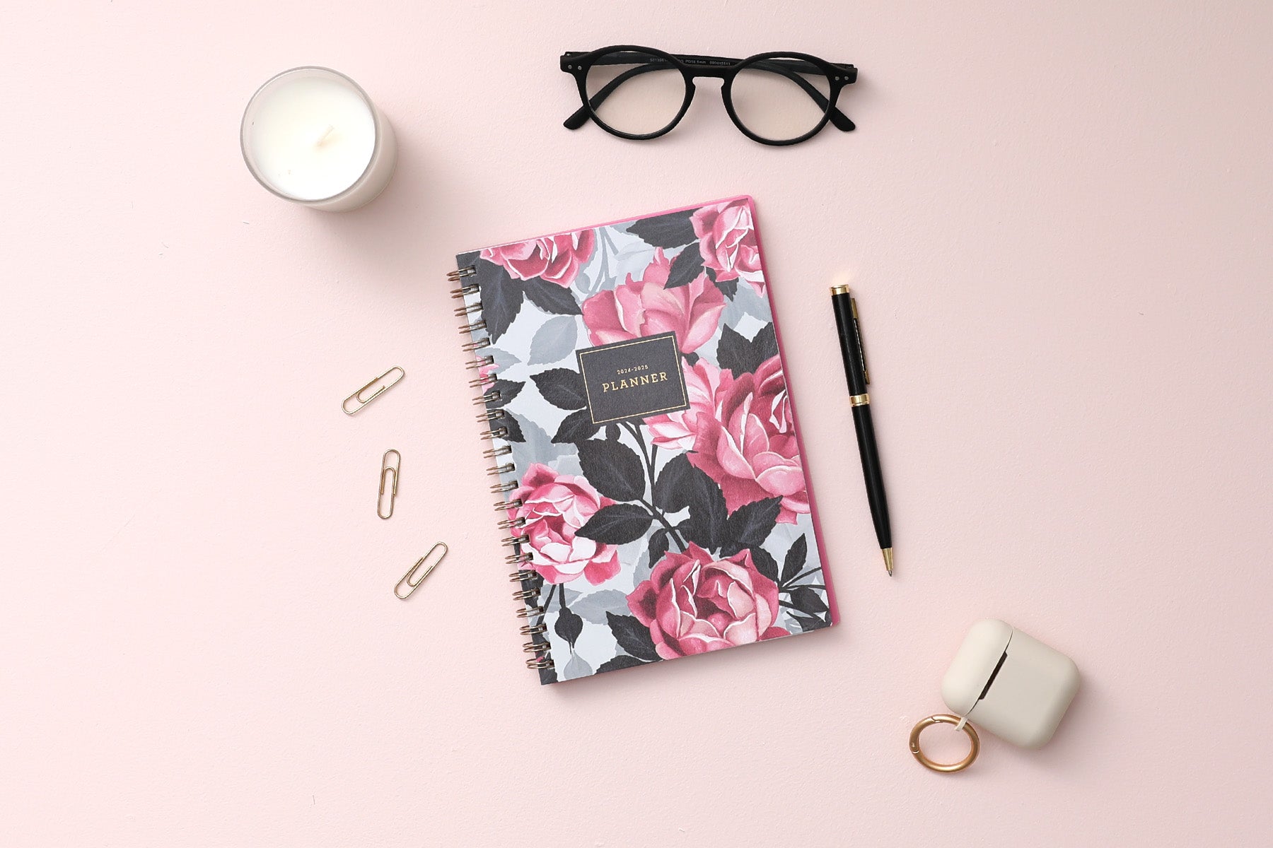 weekly monthly academic planner featuring a pink roses, shaded rose pedals, gold twin wire-o binding, and a compact 5x8 planner size, airpods, pen, paperclips, glasses, and candle on the side.