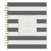 A spiral-bound planner with horizontal gray and white stripes, featuring a central gold foil label that reads &