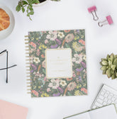 A floral spiral-bound planner or notebook lies on a white desk surrounded by styled office props, including pink binder clips, a small succulent, eyeglasses, a pastry in a bowl, and part of a keyboard. The cover has a muted multicolor floral print with a cream label panel and gold text reading &
