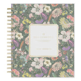 A Day Designed 2027 planner with a floral pattern cover, featuring a central cream-colored label and gold spiral binding on the left side.