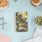 A spiral-bound 2027 floral planner lies on a light blue desk surface, surrounded by lifestyle props including a croissant on a plate, eyeglasses, a coffee cup, a succulent, and an open notebook.
