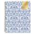 A 2027 weekly monthly planner with a white cover decorated in a blue floral and ornamental pattern, featuring a gold ribbon seal on the top right and a gold spiral binding on the left.