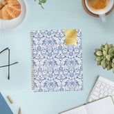 A 2027 weekly monthly planner with a white cover decorated in a blue floral and ornamental pattern, featuring a gold ribbon seal on the top right and a gold spiral binding on the left.