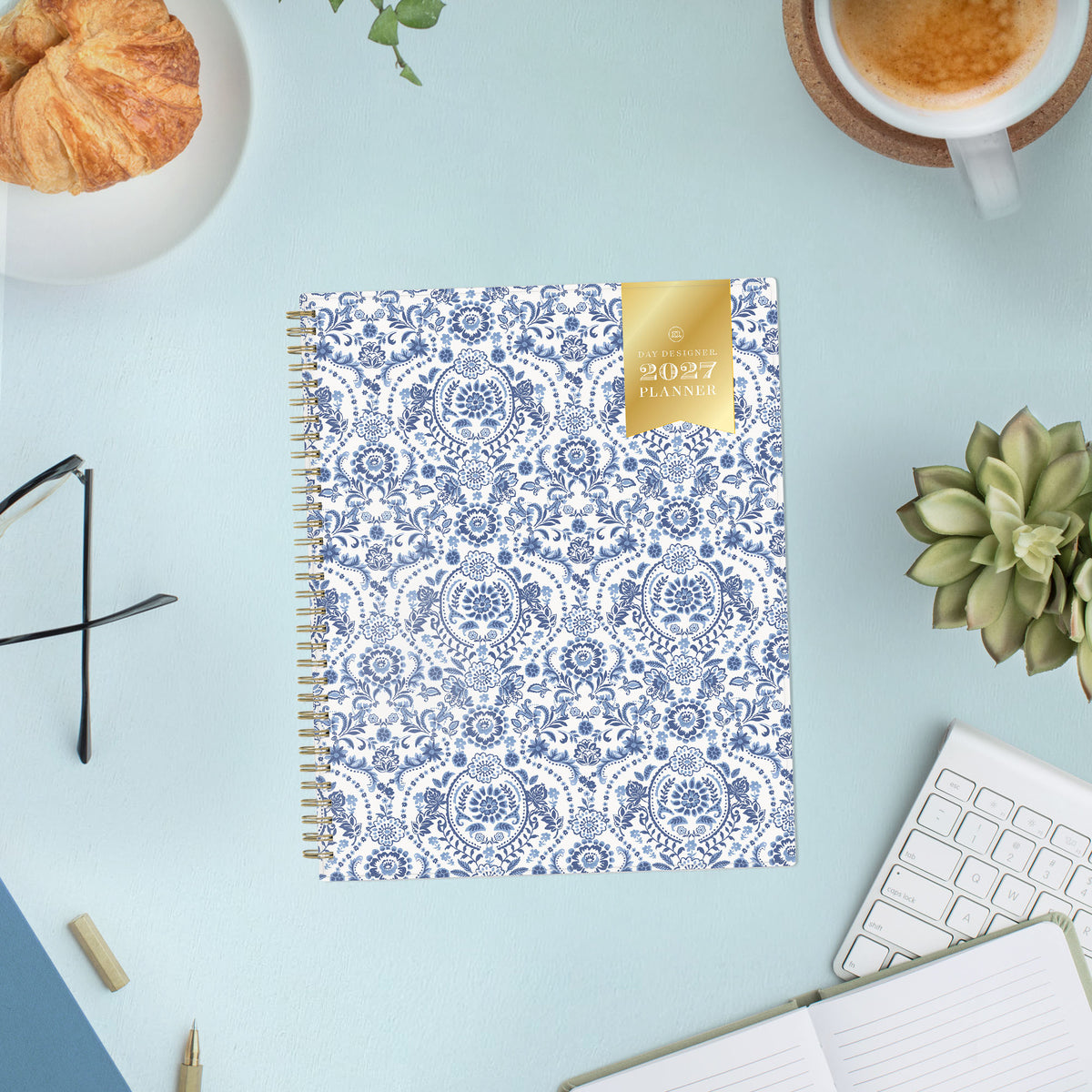 A 2027 weekly monthly planner with a white cover decorated in a blue floral and ornamental pattern, featuring a gold ribbon seal on the top right and a gold spiral binding on the left.