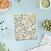 A 2027 spiral-bound weekly monthly planner with a pastel floral cover pattern featuring pink, yellow, and purple flowers with green leaves on a cream background, with a gold accent block in the top right corner.