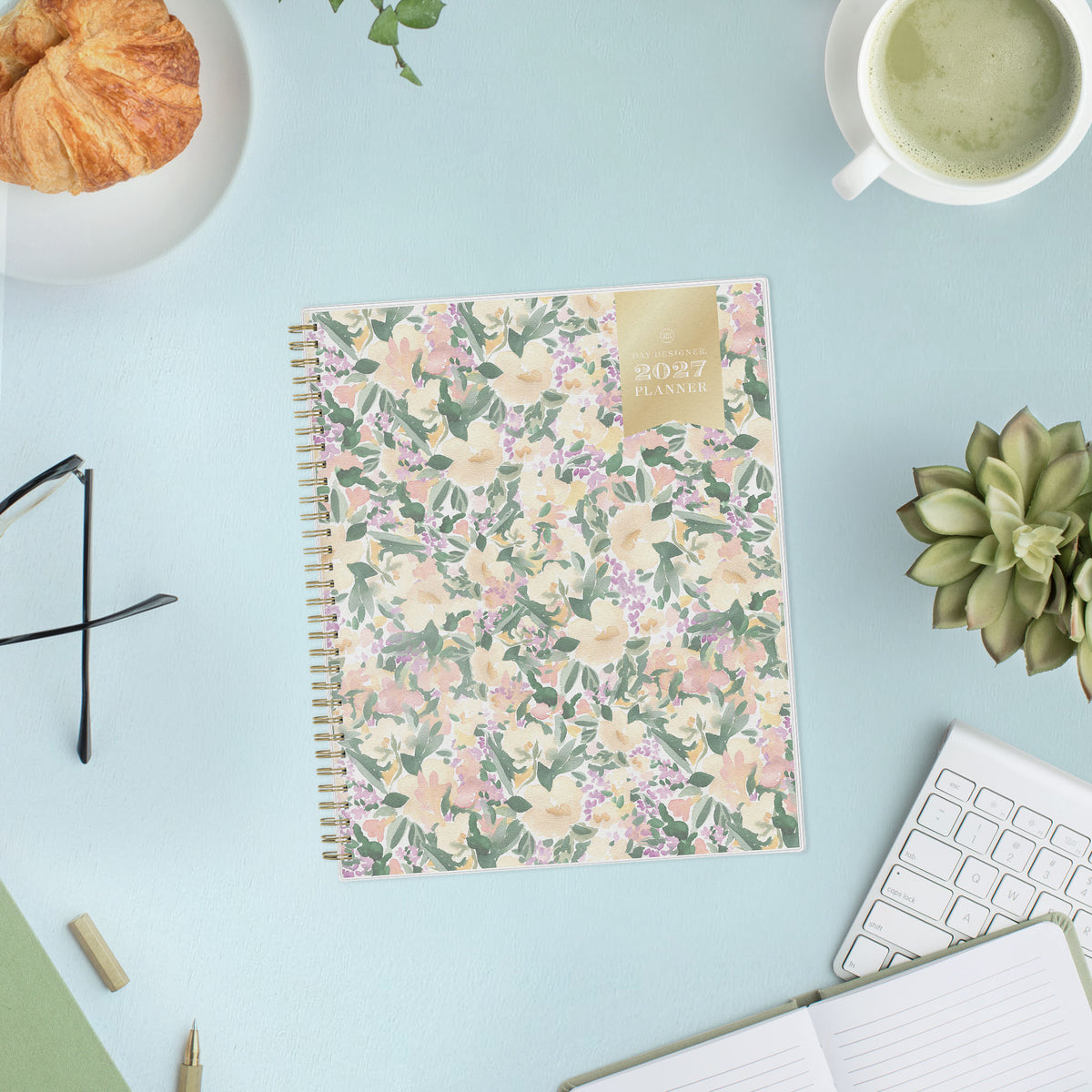 A 2027 spiral-bound weekly monthly planner with a pastel floral cover pattern featuring pink, yellow, and purple flowers with green leaves on a cream background, with a gold accent block in the top right corner.