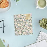A spiral-bound 2027 daily planner with a pastel floral cover pattern featuring pink, purple, and green flowers on a cream background, with a gold label on the top right.