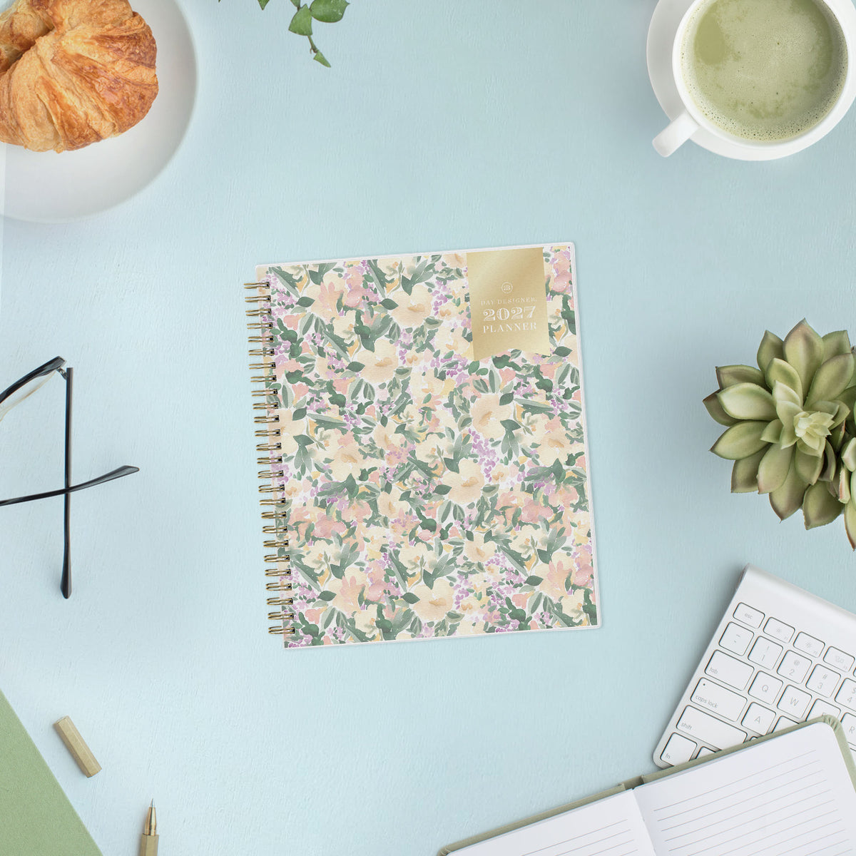 A spiral-bound 2027 daily planner with a pastel floral cover pattern featuring pink, purple, and green flowers on a cream background, with a gold label on the top right.