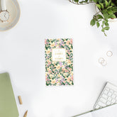 A tall daily planner with a watercolor floral cover pattern in green, pink, and cream, featuring a gold-bordered label in the center that reads &