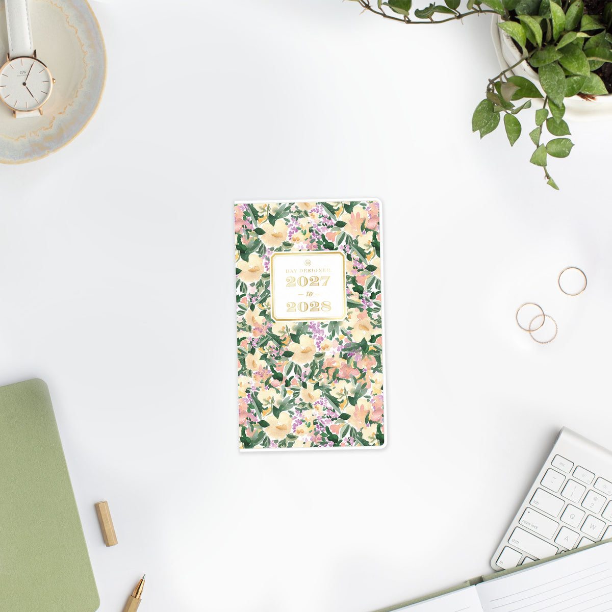 A tall daily planner with a watercolor floral cover pattern in green, pink, and cream, featuring a gold-bordered label in the center that reads &