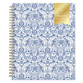 A 2027 spiral-bound weekly monthly planner with a white cover decorated in a blue floral and ornamental pattern, featuring a gold foil seal on the top right that reads &