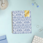 A 2027 spiral-bound weekly monthly planner with a white cover decorated in a blue floral and ornamental pattern, featuring a gold foil seal on the top right that reads &