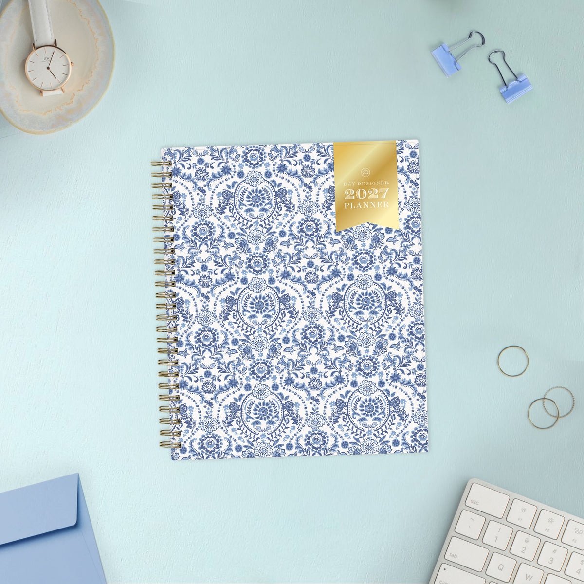 A 2027 spiral-bound weekly monthly planner with a white cover decorated in a blue floral and ornamental pattern, featuring a gold foil seal on the top right that reads &