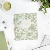 A spiral-bound planner with a green botanical toile pattern and styled on a white desk with a coffee cup, keyboard, plant, and other desk accessories.
