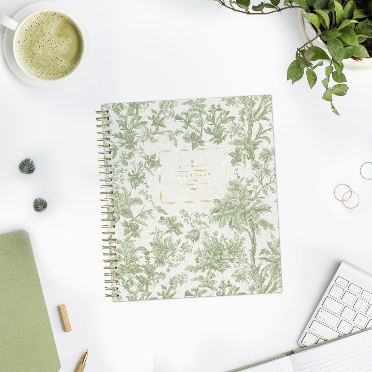 A spiral-bound planner with a green botanical toile pattern and styled on a white desk with a coffee cup, keyboard, plant, and other desk accessories.