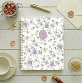 A spiral-bound non-dated daily planner with a white cover decorated with a repeating pattern of purple roses and green leaves, featuring a round lavender sticker with gold foil text that reads &