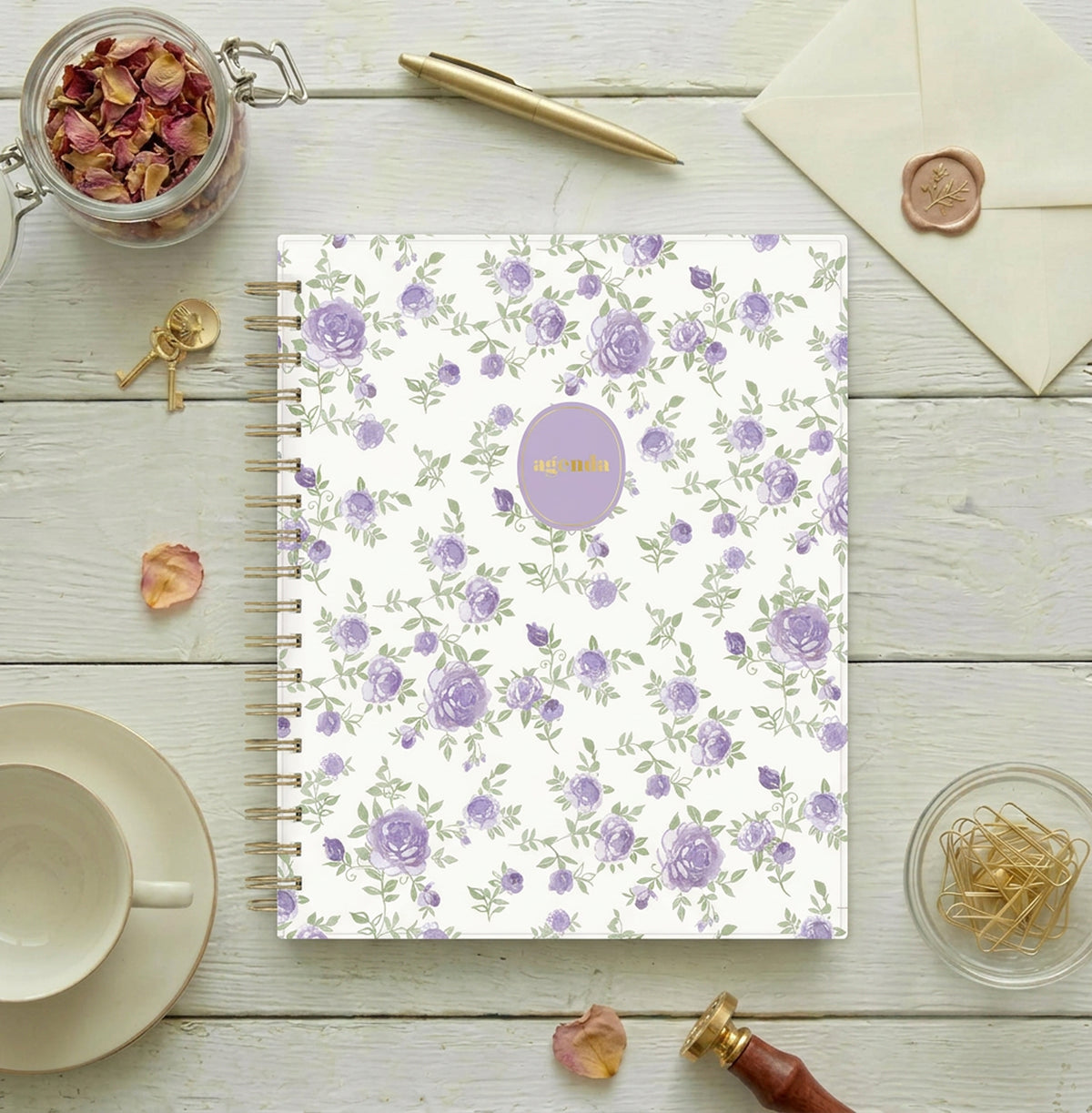 A spiral-bound non-dated daily planner with a white cover decorated with a repeating pattern of purple roses and green leaves, featuring a round lavender sticker with gold foil text that reads &