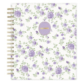 A spiral-bound non-dated daily planner with a white cover decorated with a repeating pattern of purple roses and green leaves, featuring a round lavender sticker with gold foil text that reads &