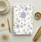 A spiral-bound planner agenda with a white cover decorated with a purple rose and green leaf pattern, featuring a lavender oval label with gold foil text that reads &