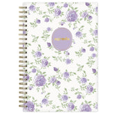 A spiral-bound planner agenda with a white cover decorated with a purple rose and green leaf pattern, featuring a lavender oval label with gold foil text that reads &