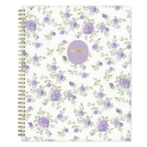 A spiral-bound weekly monthly planner with a white cover decorated with a repeating pattern of purple roses and green leaves. A lavender circular sticker with gold foil text reading &