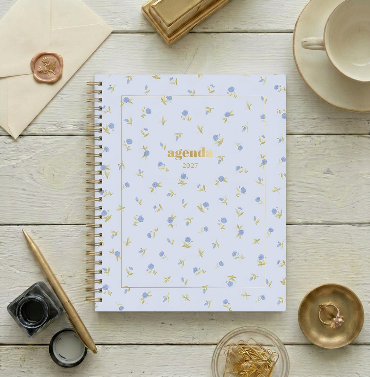 A spiral-bound 2027 agenda with a light blue cover decorated with a delicate pattern of small blue flowers and yellow-green leaves. A circular blue label in the center features the word &