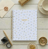 A spiral-bound 2027 agenda with a light blue cover decorated with a delicate pattern of small blue flowers and yellow-green leaves. A circular blue label in the center features the word &