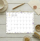 A spiral-bound monthly calendar page for January 2027, featuring a white background with light purple floral borders and gold accents, including a gold spiral binding and a gold clip at the top.