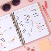 Open planner with notes and to-do list on a pink surface with sunglasses, pens, and clips, featuring a weekly spread