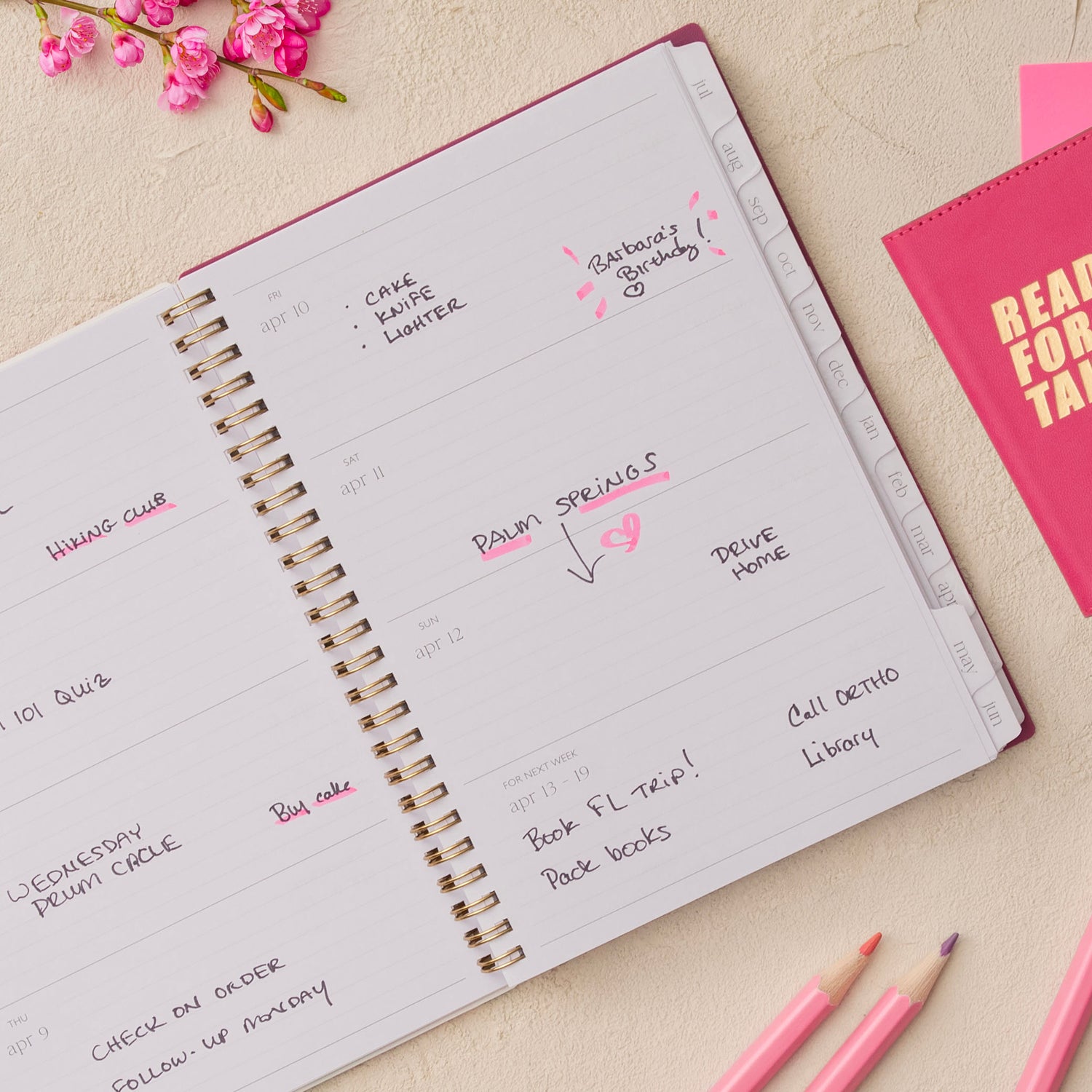 Open spiral-bound planner with handwritten notes on a light surface with pink flowers and stationery items., lined writing space white monthly tabs
