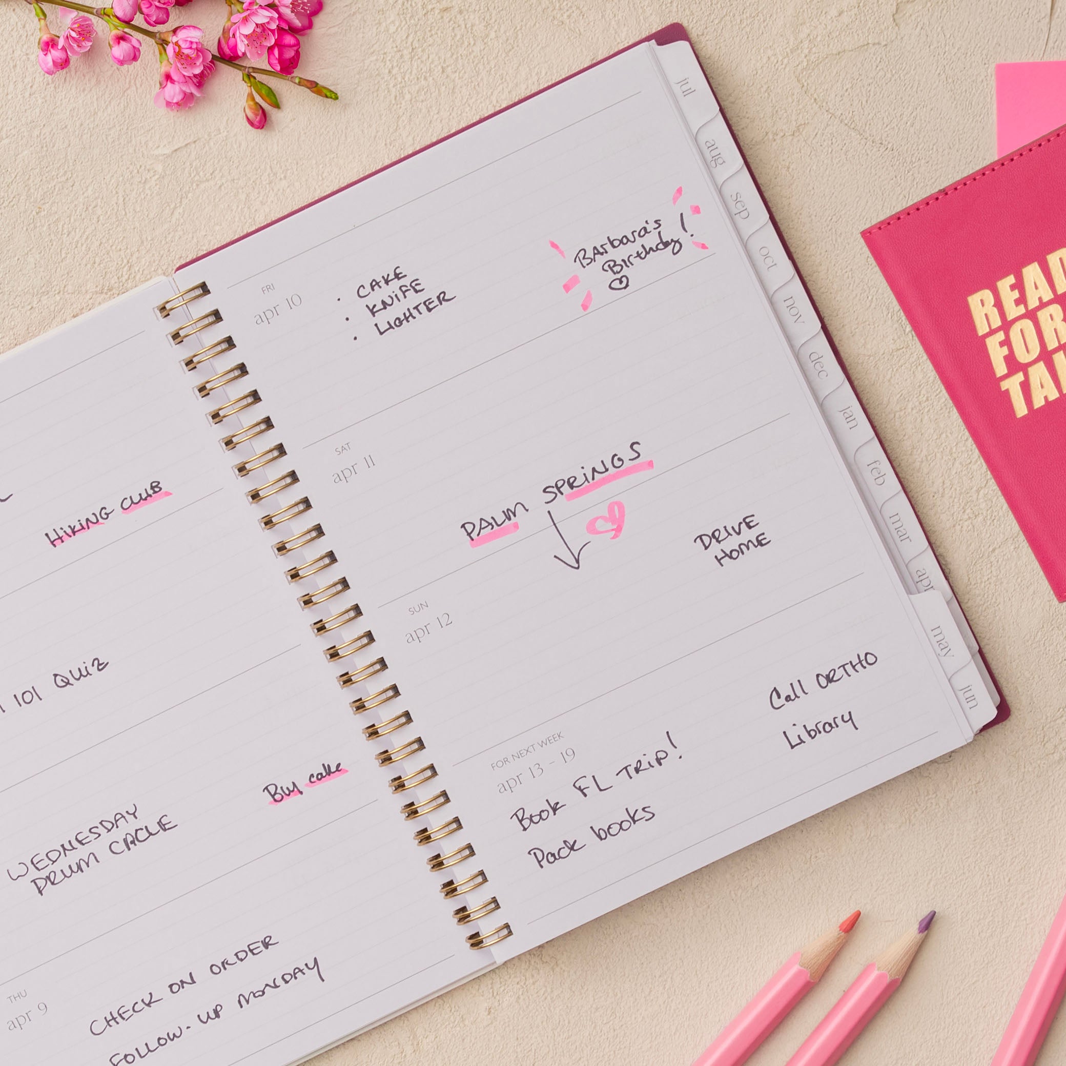 Open spiral-bound planner with handwritten notes on a light surface with pink flowers and stationery items., lined writing space white monthly tabs