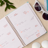 Open planner with to-do list on a sandy surface with sunglasses and shells, lined writing space and white monthly tabs